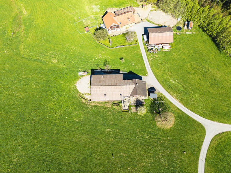 Aerial view of a rural property with multiple buildings, including a main house, outbuildings, and a parking area, surrounded by green fields and trees.