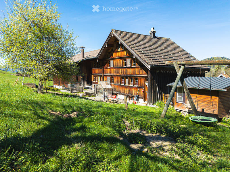 A two-story wooden house with a sloped roof, multiple windows, and a balcony, surrounded by a grassy yard and trees.