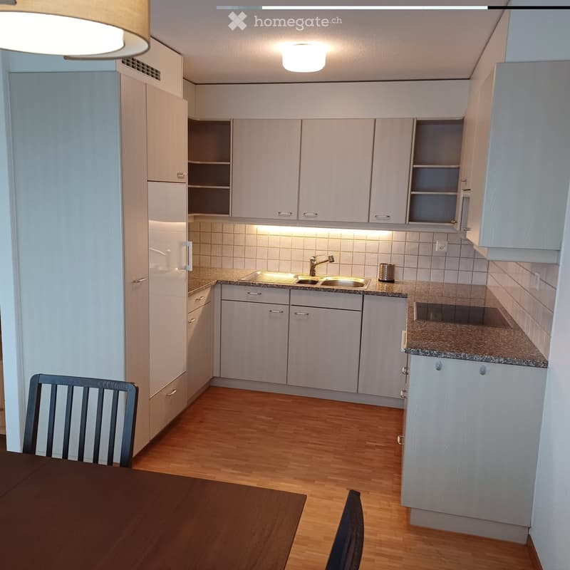 Furnished Room. Modern kitchen with light-colored cabinets, granite countertops, and a sink. A wooden dining table and chairs are visible in the foreground.