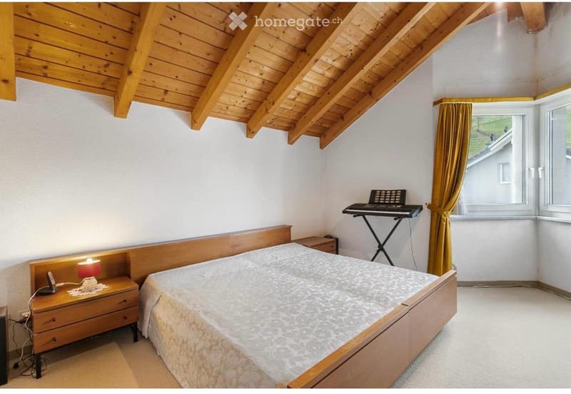 Cozy bedroom with wooden ceiling beams, a double bed with patterned bedding, a nightstand with a lamp, and a small keyboard on a stand near the window.