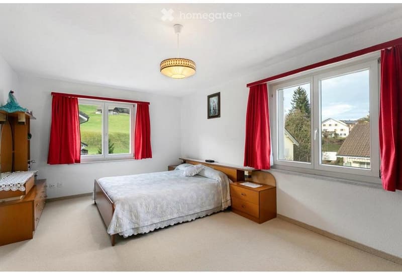 Bright bedroom with large windows offering a view of a green landscape, featuring a wooden bed, nightstand, wardrobe, and decorative picture frame.