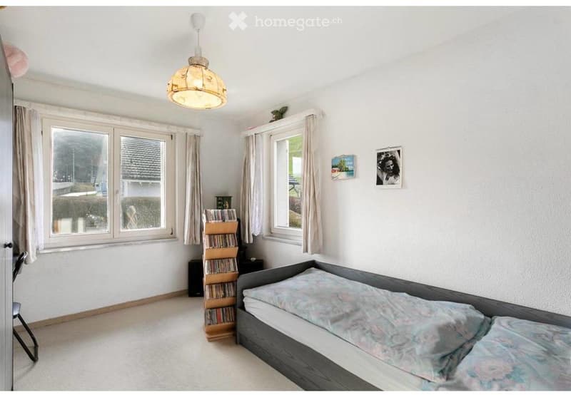 A well-lit bedroom with a grey bed, a wooden bookcase, and large windows with curtains. The room features a hanging lamp and several picture frames on the wall.