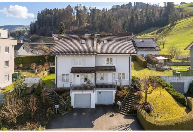 Two-story white house with a tiled roof, garage, and balcony overlooking a green, hilly landscape with neighboring houses and trees.