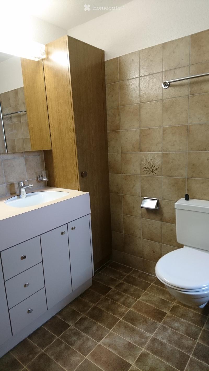 Modern bathroom with beige tiles, a white sink with a chrome faucet, a wooden cabinet, and a white toilet. The walls are tiled in a warm beige color, and the floor is covered with brown tiles.