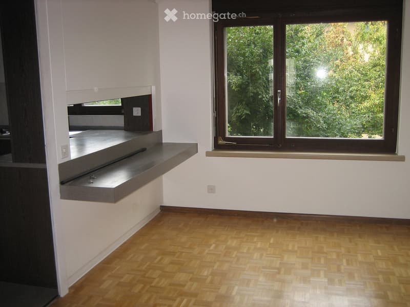 Empty room with wooden flooring, white walls, a large window with a view of greenery, and a built-in shelf with a small black object on it.