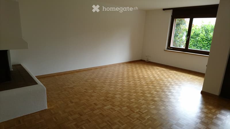 Empty room with wooden herringbone flooring, white walls, and a large window with a view of greenery.