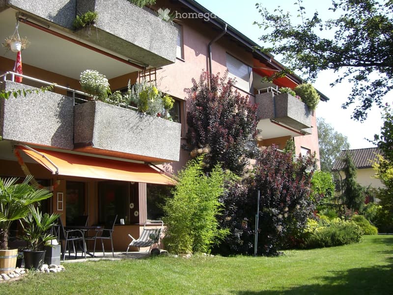 Modern two-story house with balconies, greenery, and outdoor seating area.