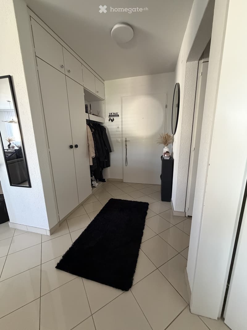 Clean hallway with tiled floor, white walls, built-in closet with hanging clothes, small black rug, and decorative items on a side table.