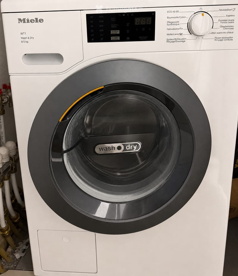 A modern white Miele washing machine with a digital control panel and a clear front door, featuring a 'wash & dry' label.