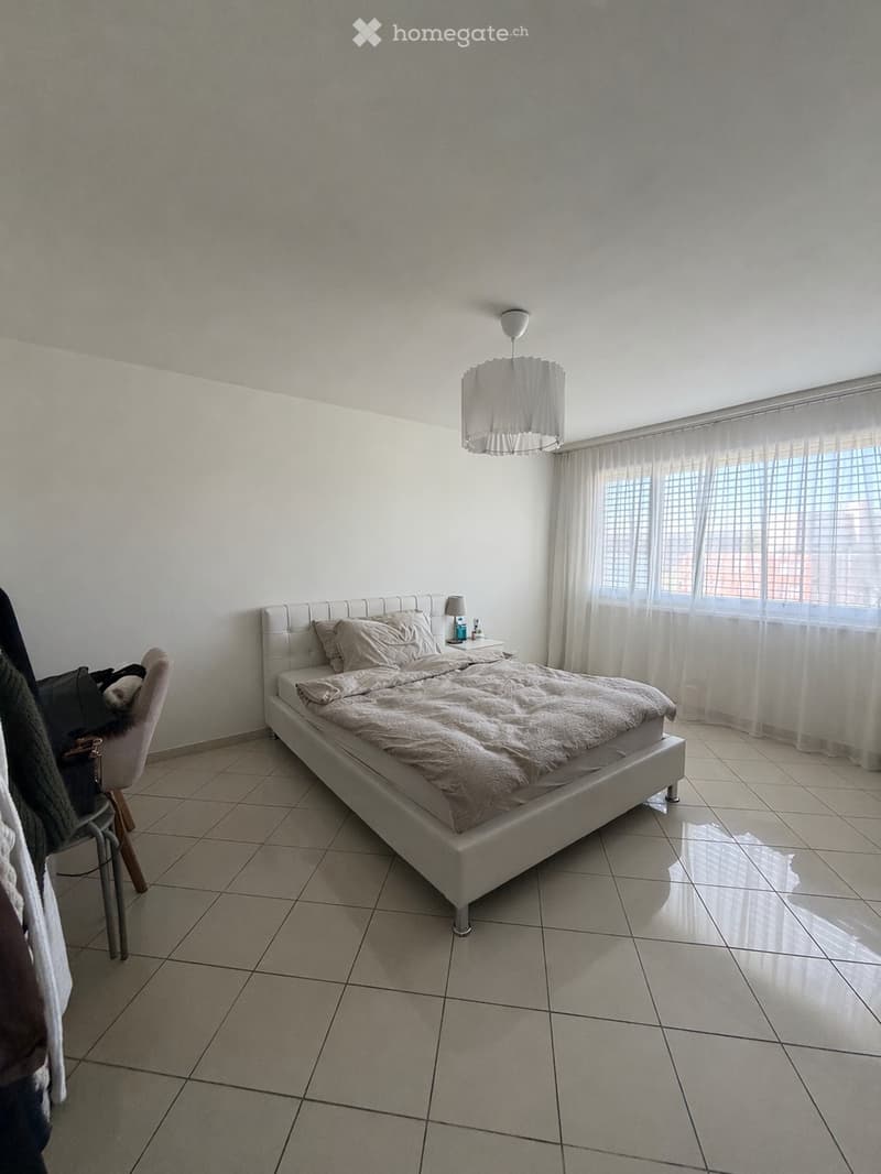 A modern bedroom with a large bed, a nightstand with a lamp, a chair, and large windows with sheer curtains.