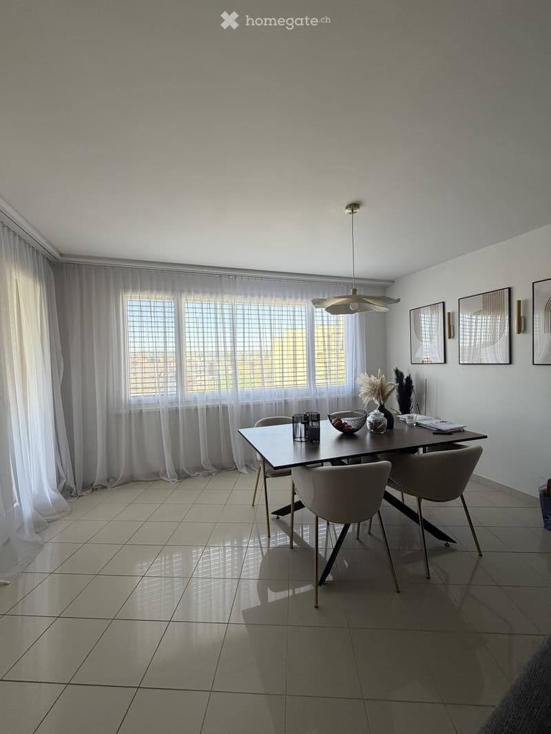 Modern dining room with a sleek dining table, four chairs, decorative items, and large windows with sheer curtains.
