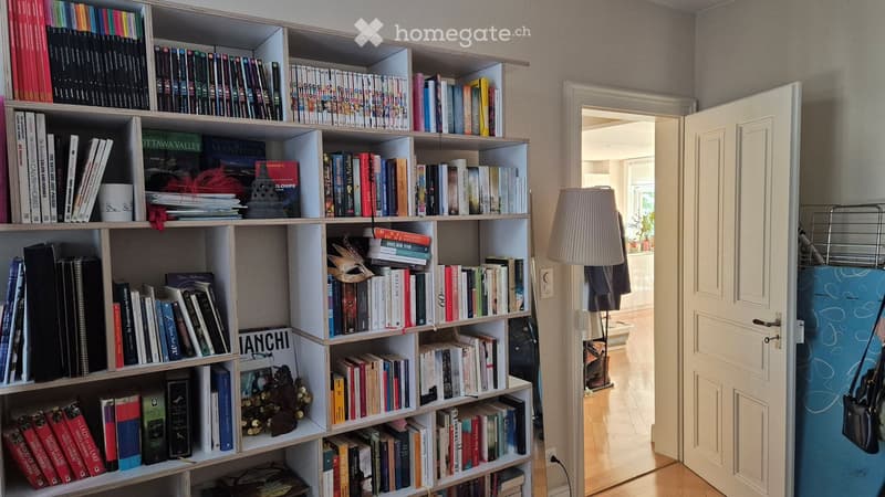 A cozy room with a large wooden bookshelf filled with books, a table lamp, and a cup on the shelf. An open door leads to another room with a visible refrigerator.
