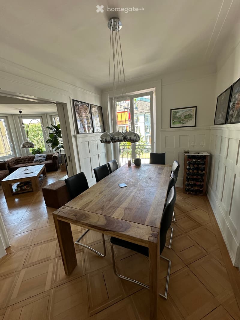 Modern dining area with a large wooden dining table surrounded by black chairs, a sofa, coffee table, and a wine rack. Large windows and a view of the balcony are visible.