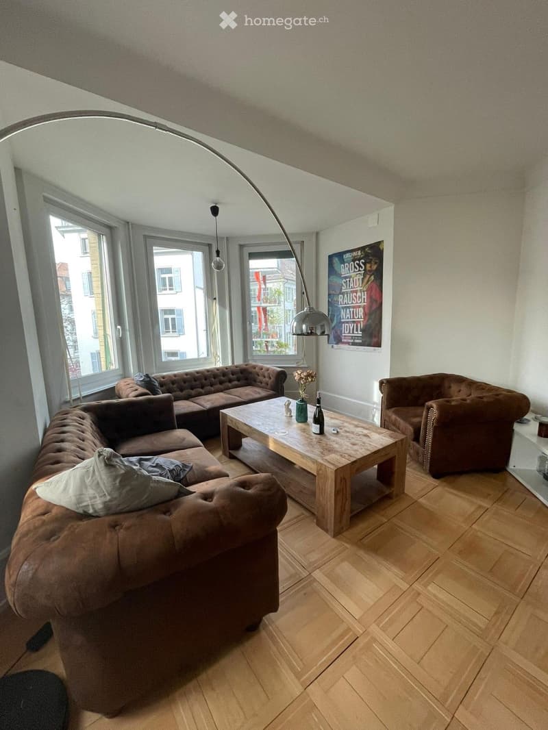 Modern living room with wooden flooring, large windows, a brown leather sofa, a wooden coffee table, and an armchair. A floor lamp and a vase with flowers are also present.
