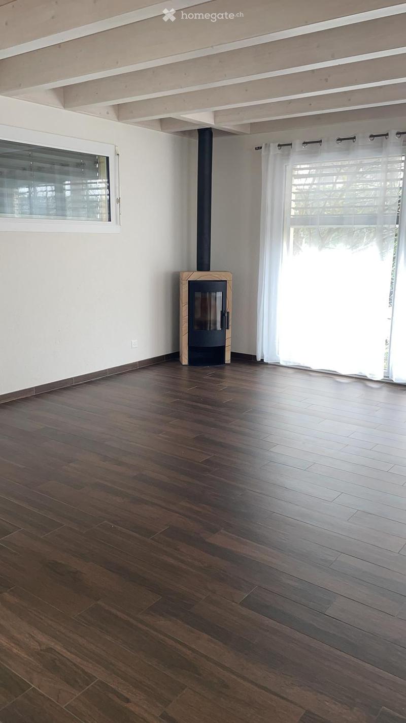 Empty room with wooden flooring, white walls, large windows with white curtains, and a modern fireplace with a chimney.