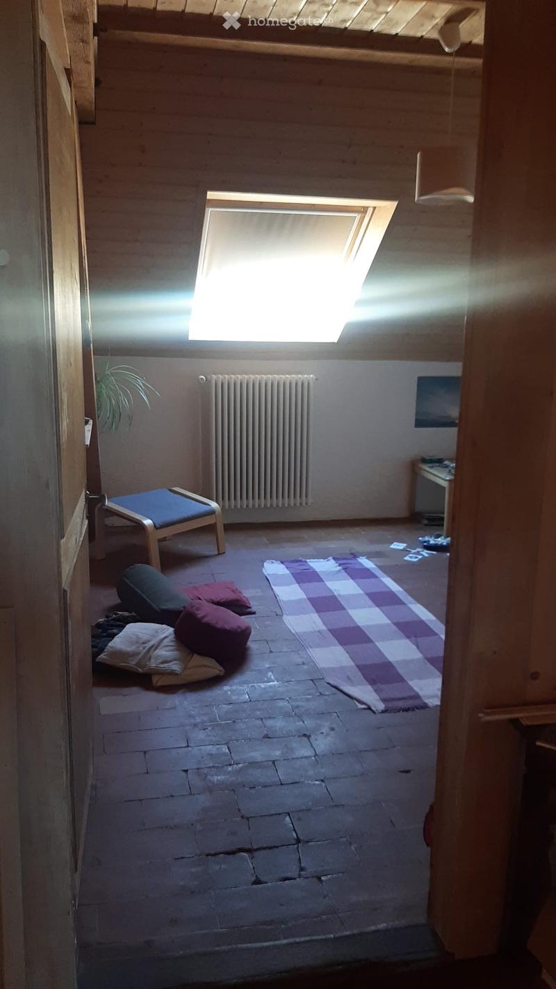 Loft-style room with wooden beams, skylight window, stone floor, and minimal furnishings including a stool, pillows, and a rug.