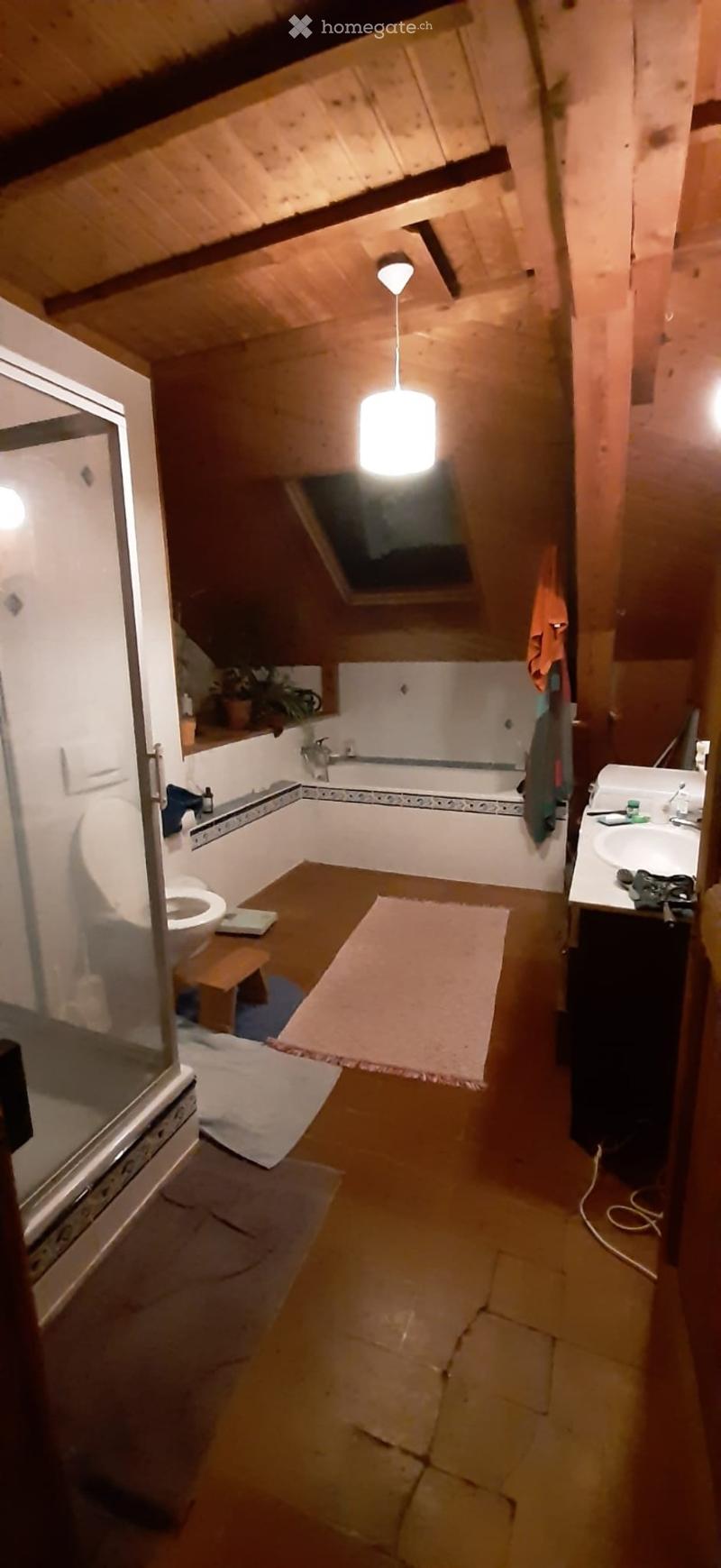 Bathroom with wooden ceiling and walls, featuring a toilet, bathtub, sink, small stool, mirror, and a hanging light fixture. A small plant is placed on the windowsill, and towels are visible.