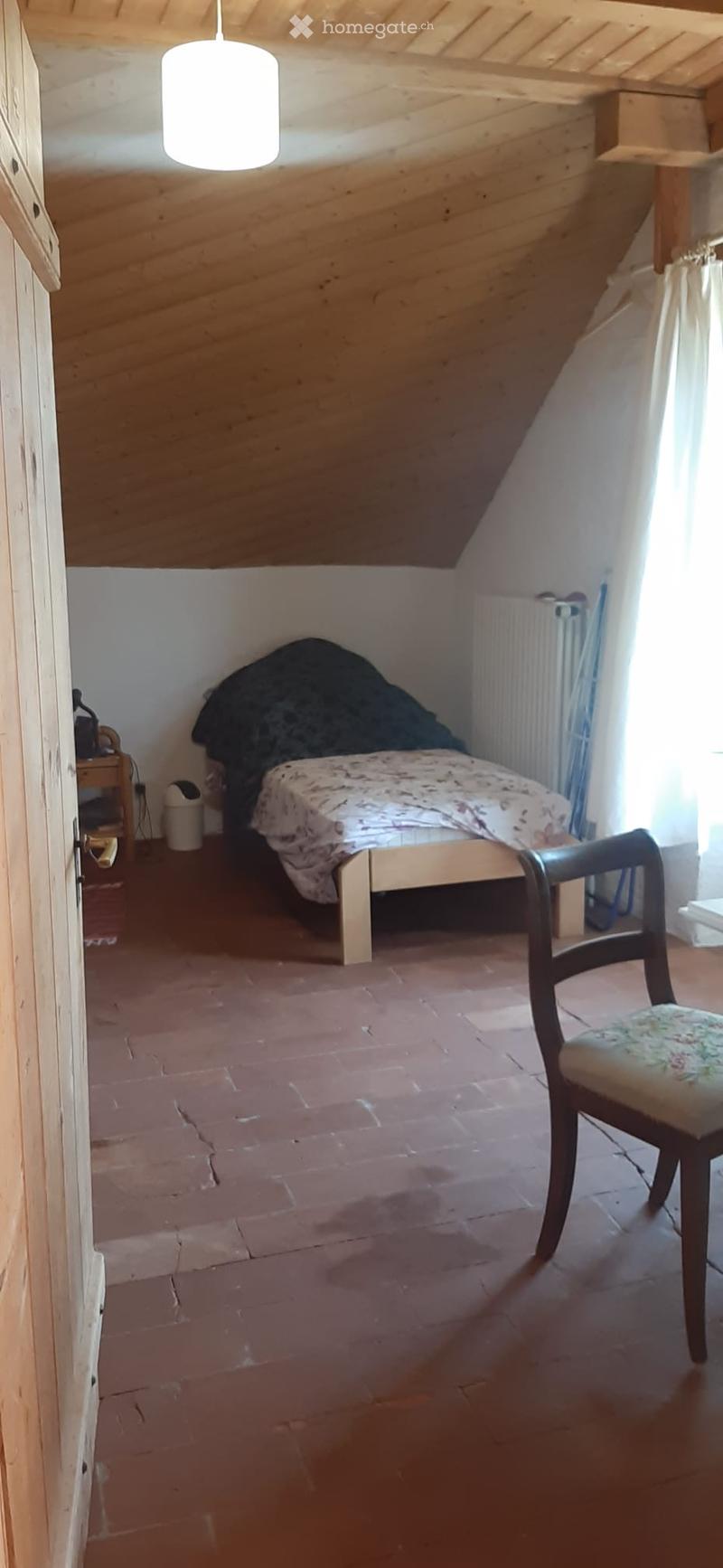 Cozy attic bedroom with wooden slanted ceiling, terracotta floor, single bed with floral bedding, wooden nightstand, and a wooden chair.