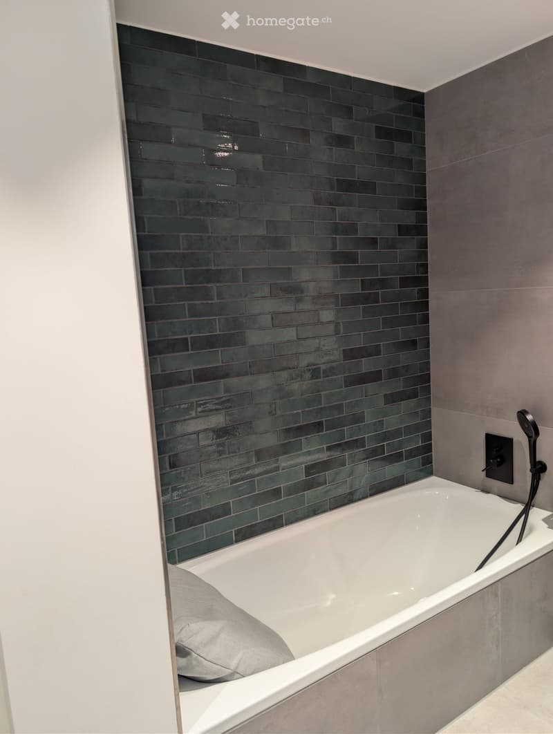 Modern bathroom with a white bathtub, dark green tiled accent wall, and a showerhead mounted on the wall.