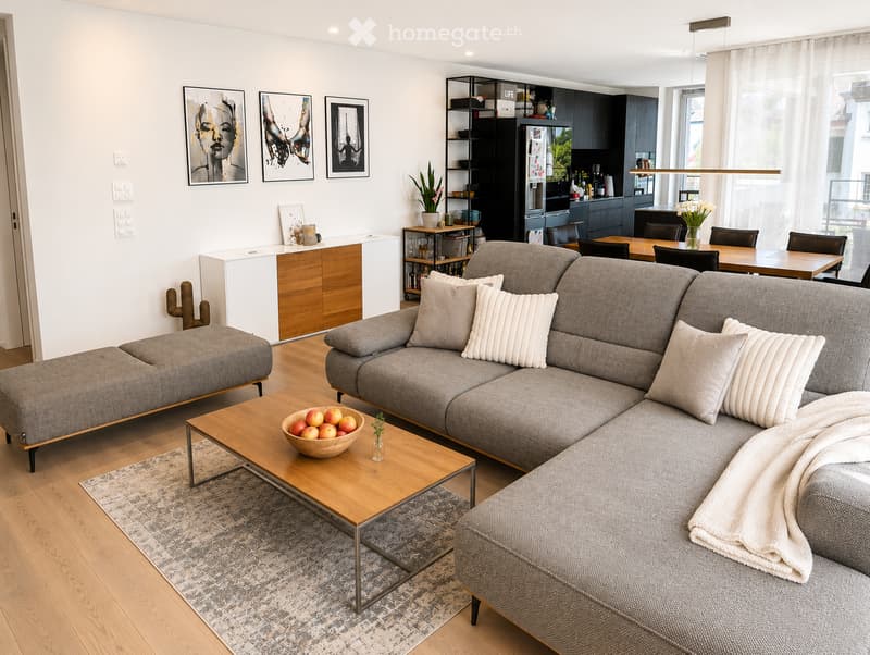 Modern living room with grey sectional sofa, wooden coffee table, ottoman, and dining area visible in the background.