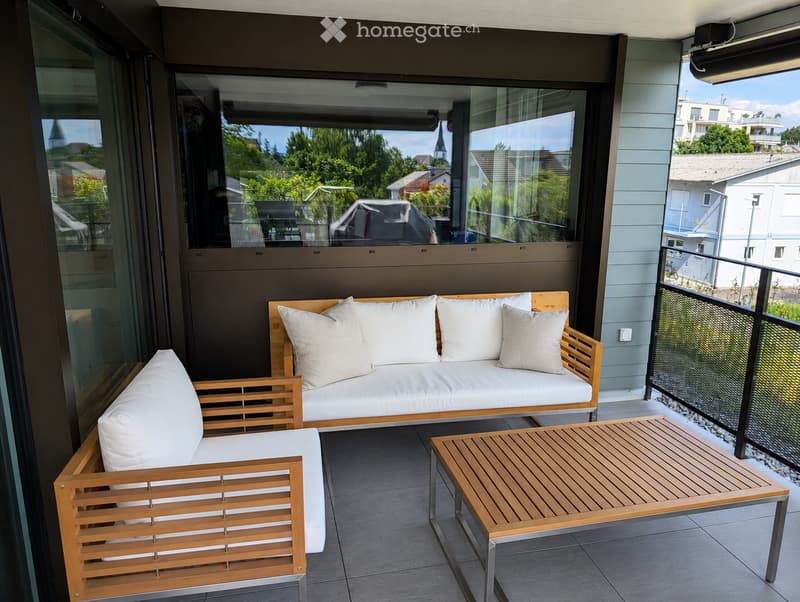 Modern balcony with a wooden sofa, two wooden chairs, and a wooden coffee table, overlooking a green landscape.