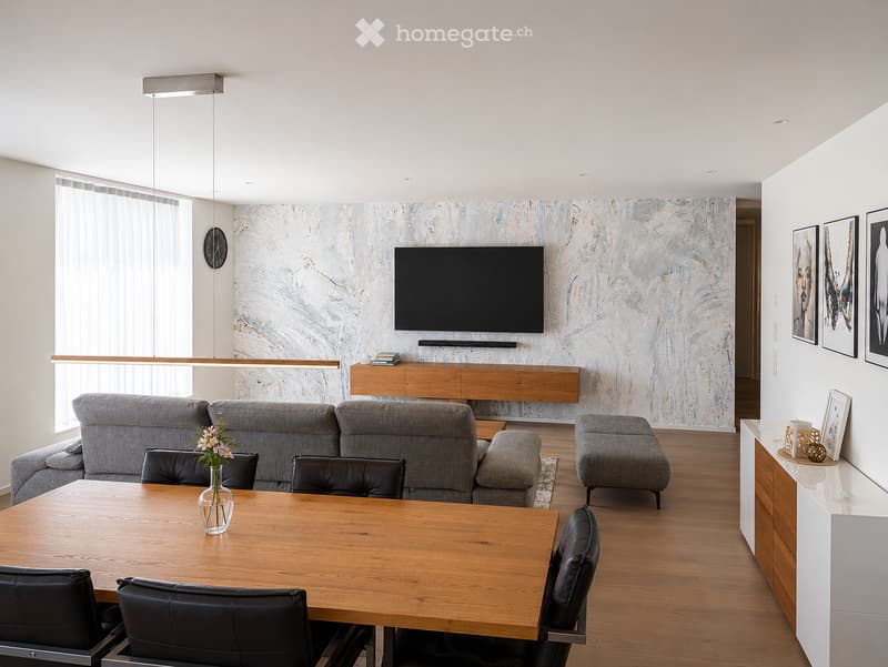 Modern living room with a grey sectional sofa, wooden coffee table, dining table with black chairs, TV mounted on a decorative wall, and artwork displayed on the walls.