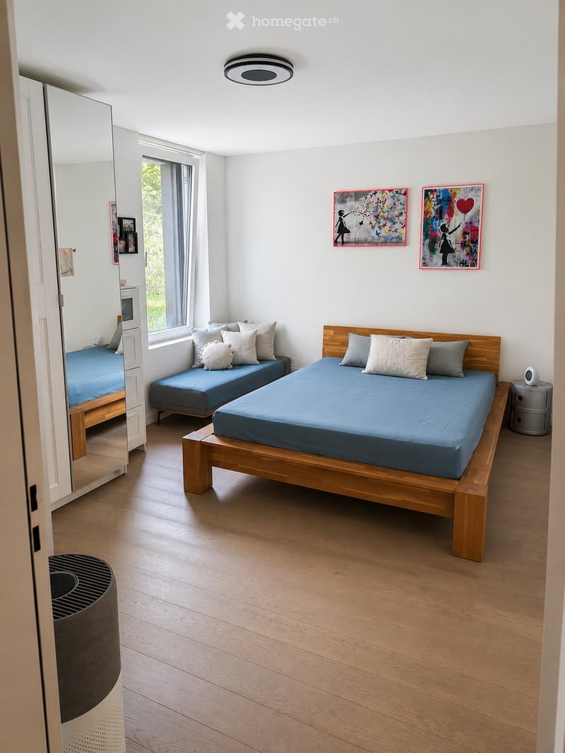 Bright bedroom with wooden flooring, two beds with blue bedding, nightstand, mirror, and artwork on the walls.