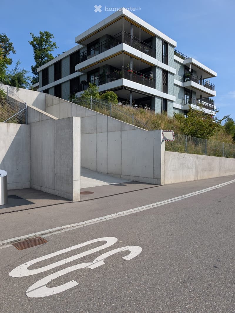 Modern multi-story residential building with balconies, surrounded by a concrete barrier and greenery.