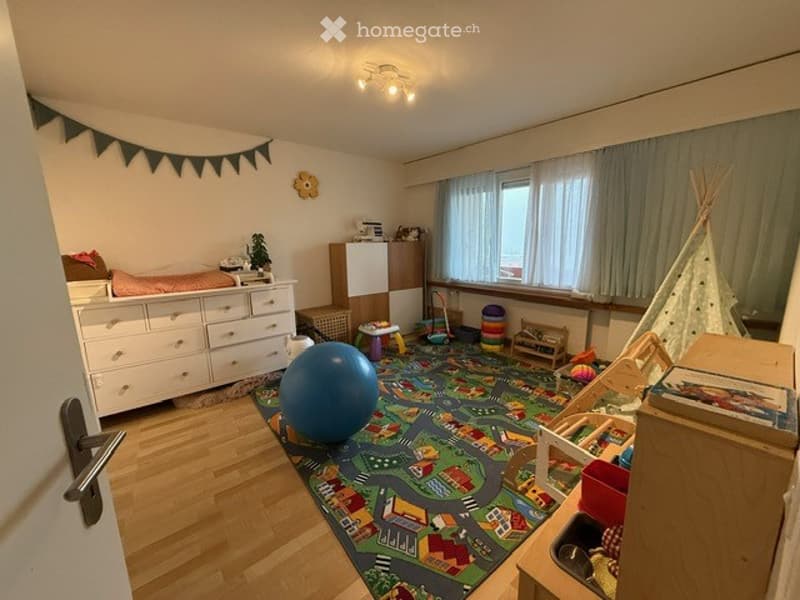 A well-lit children's room with a colorful play mat, a large blue balloon, a small teepee tent, and various toys scattered around. The room features a white dresser, a small table, and a window with curtains.