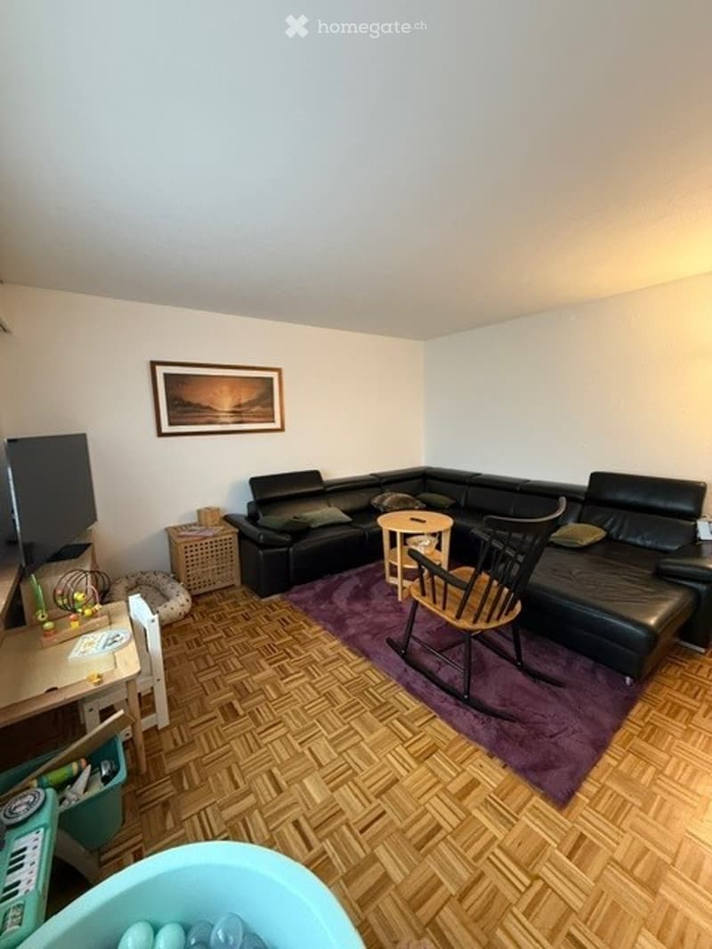 Living room with a black sectional sofa, a small round coffee table, a wooden rocking chair, a small table, a picture frame on the wall, and a small keyboard on a table.