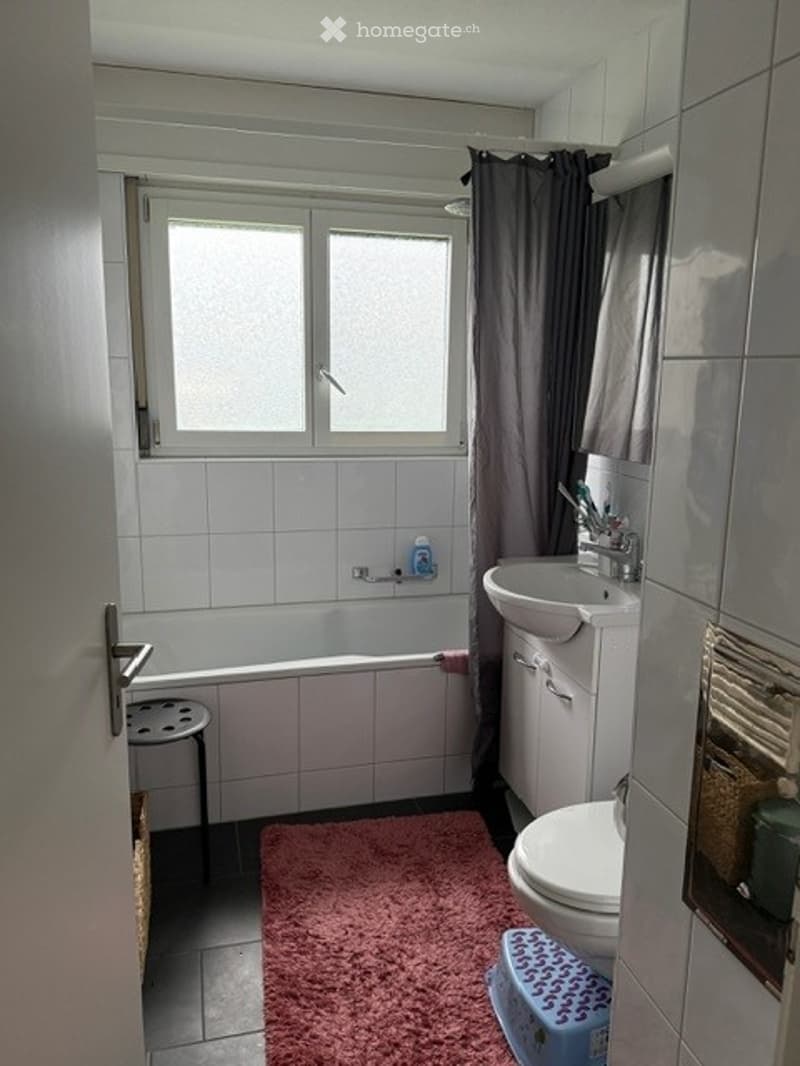 Bathroom with a bathtub, sink, toilet, and a small stool. The room features white tiled walls, a window with a curtain, and a red bath mat on the floor.