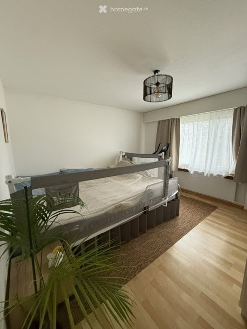 Cozy bedroom with a wooden bed, nightstand, and a hanging lamp. The room features light-colored walls, wooden flooring, and a large window with curtains.