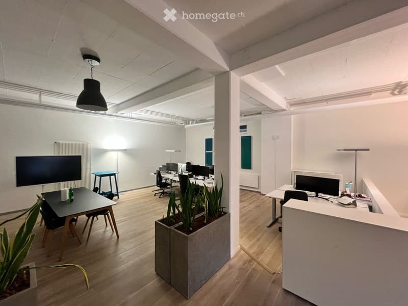 Modern office space with wooden flooring, desks, chairs, screens, and plants in large planters.