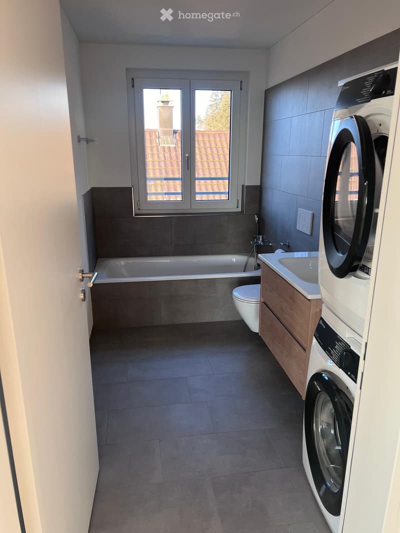 Modern bathroom with a bathtub, toilet, sink, and washing machine. Features tiled walls and a window overlooking a roof.