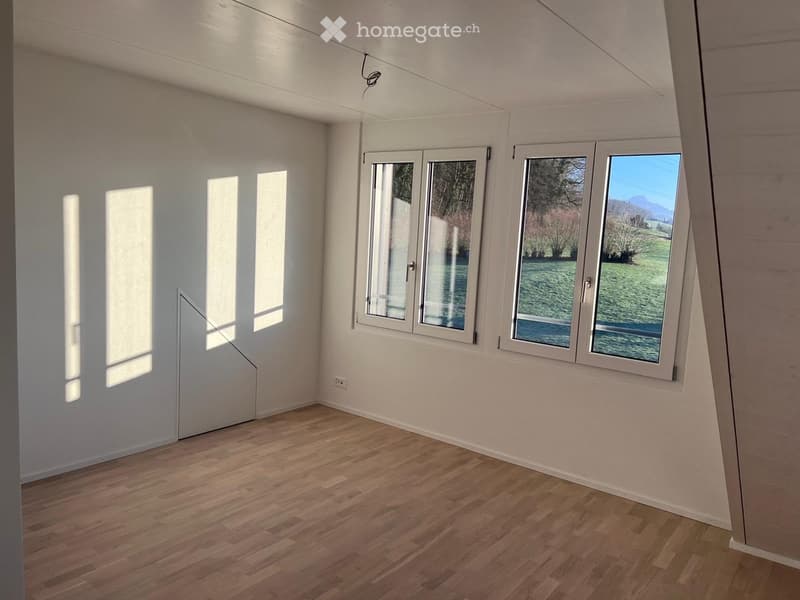 Empty room with wooden flooring, white walls, and large windows offering a view of a green landscape.
