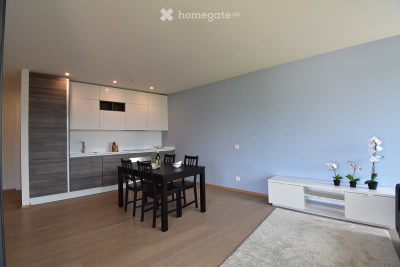 Modern kitchen and dining area with white cabinets, dark grey lower units, a black dining table with four chairs, and a white shelf with decorative flowers.