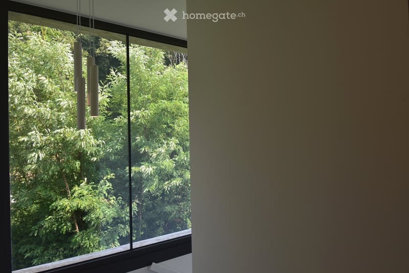 An unfurnished room with large windows offering a view of lush greenery outside. The room features a modern design with a visible light fixture hanging from the ceiling.