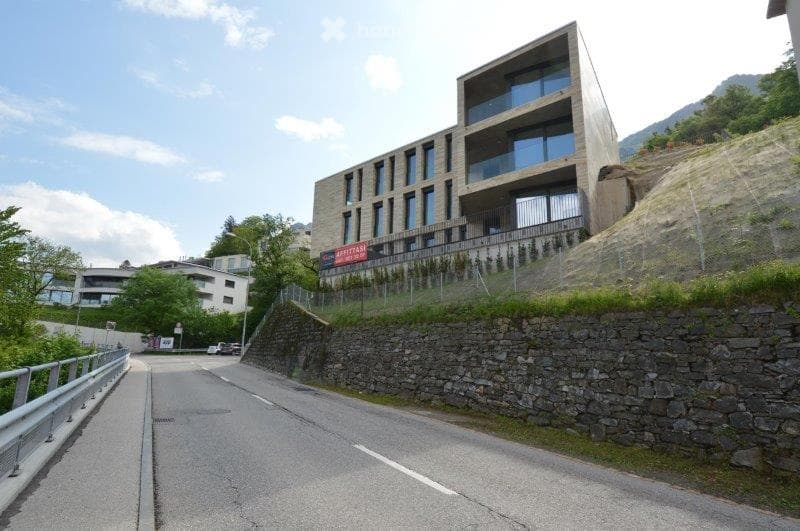 Modern multi-story building with large glass windows and balconies, situated on a hillside with a stone retaining wall and greenery.