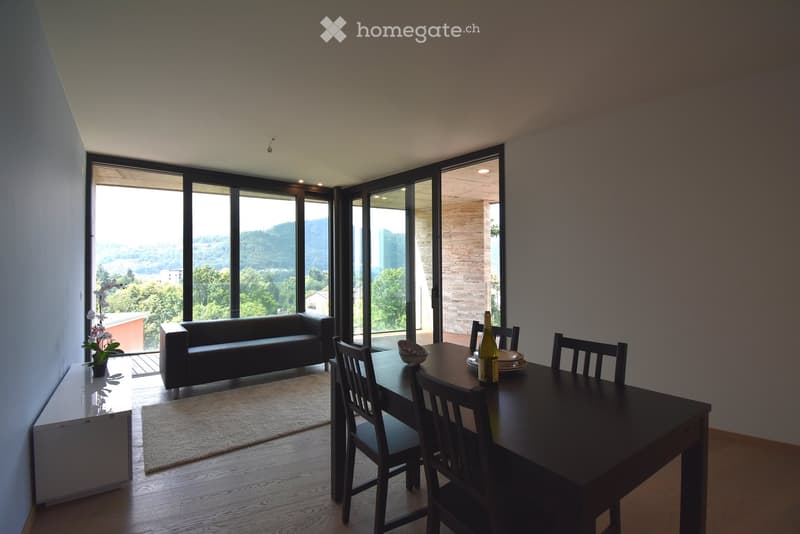 Modern living and dining area with large windows offering a scenic view, featuring a sofa, dining table, chairs, and a wine bottle on the table.