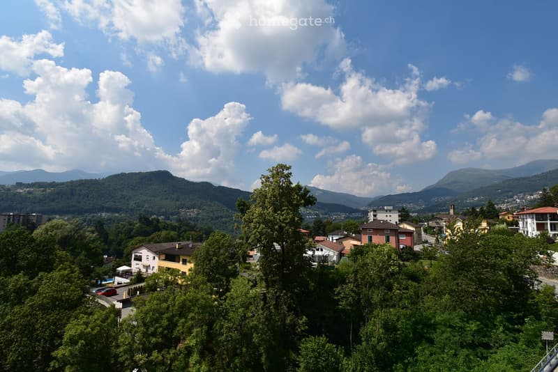 A scenic view of a town nestled in a valley surrounded by lush green trees and distant mountains under a partly cloudy sky.