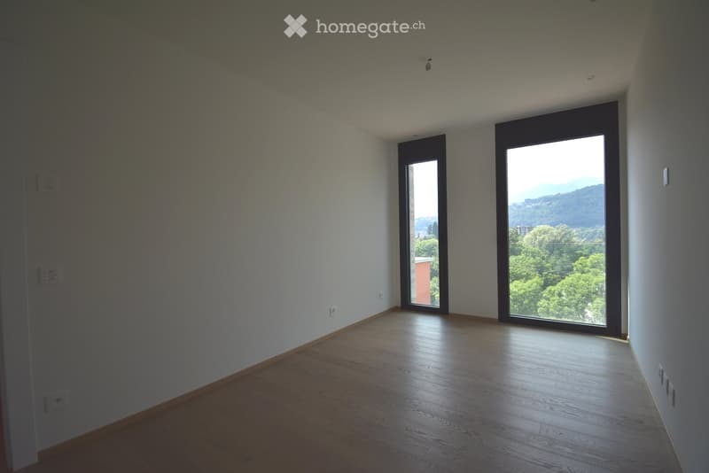 Empty room with wooden flooring, white walls, and large windows offering a scenic view of greenery and hills.
