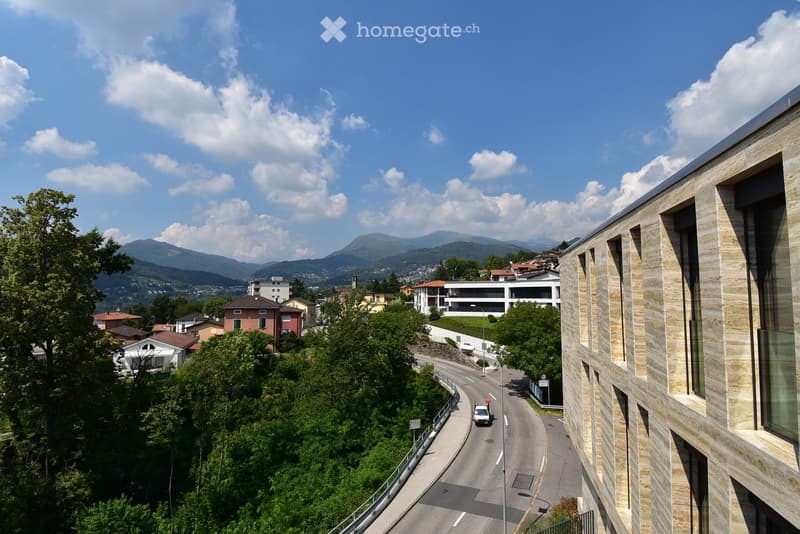 View of a modern building with stone facade, overlooking a winding road surrounded by greenery and distant mountains.