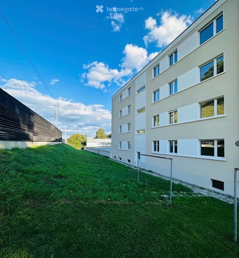 Modern multi-story building with white facade and large windows, surrounded by a grassy area and a wooden fence.