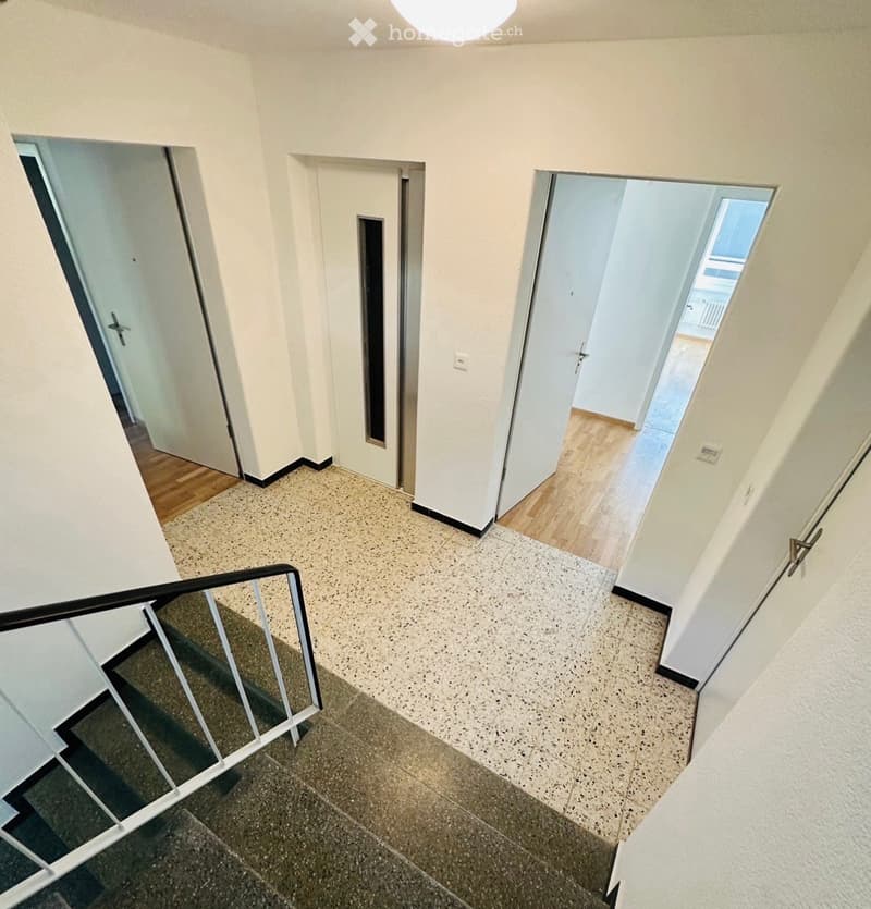 A clean, modern hallway with white walls, tiled floor, and a staircase leading upwards. Doors open to adjacent rooms, one with wooden flooring visible.