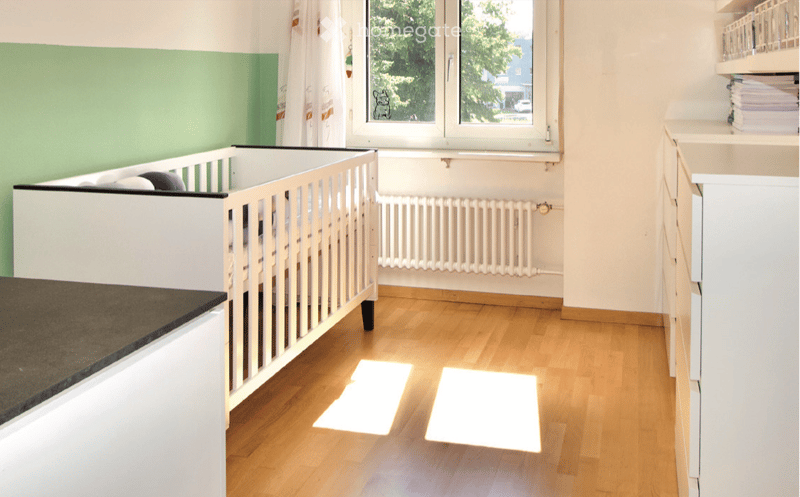 A well-lit room with a wooden floor, a white crib, a radiator, a white cabinet, and a shelf with books and other items.