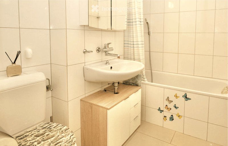 Modern bathroom with white tiles, a sink with a chrome faucet, a toilet with a marble-patterned seat cover, a small wooden cabinet, a bathtub with butterfly decals, and a shower curtain with stripes.