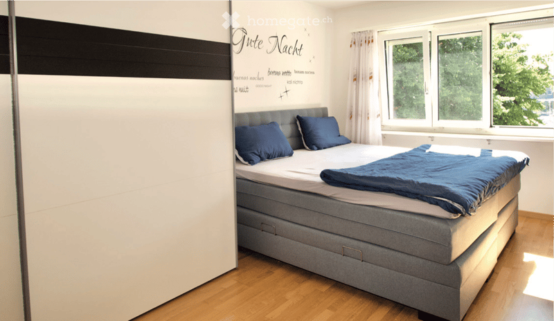 A modern bedroom with a large bed, blue pillows, a blue blanket, and a grey wardrobe. The room has wooden flooring and large windows with white curtains.