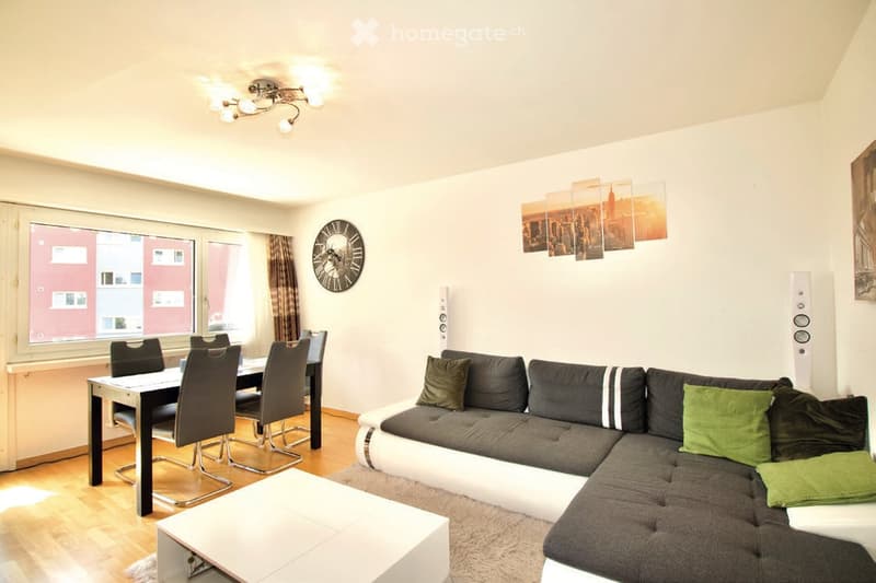 Modern living and dining area with a grey sectional sofa, dining table with chairs, coffee table, wall clock, and speakers.
