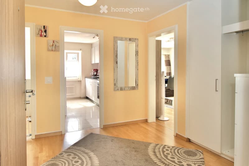 Bright hallway with wooden flooring, a large mirror on the wall, and a patterned rug on the floor. The hallway leads to a kitchen and another room with white cabinets and a stand mixer.