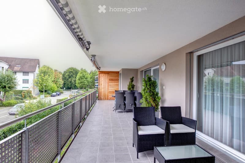 Balcony with wicker chairs, glass table, and a view of the street and greenery.